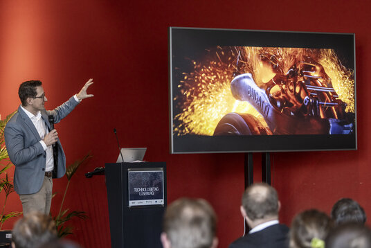 Präsentation von 3D-gedruckten Bauteilen für die Automobilindustrie | © Julia Crawley Photography Ein Mann mit Mikrofon steht auf der Bühne des Technologietags Lüneburg und zeigt auf eine große Leinwand, auf der ein funkenstiebendes 3D-gedrucktes Bugatti-Bauteil zu sehen ist.