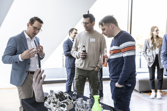 Technologiedemonstration auf dem Technologietag Lüneburg | © Julia Crawley Photography Ein Mann in hellblauem Sakko erklärt zwei Besuchern die ausgestellten 3D-gedruckten Objekte auf einem schwarzen Tisch. Die Besucher hören aufmerksam zu, einer hält eine Cola-Flasche in der Hand.
