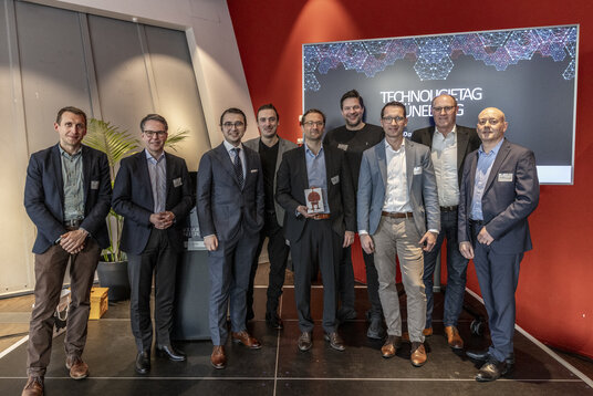 Eine Gruppe von neun Personen, bestehend aus Referenten und Veranstaltern, steht vor einer Präsentationswand mit der Aufschrift "Technologietag Lüneburg". Einer der Teilnehmer hält eine Trophäe in den Händen.