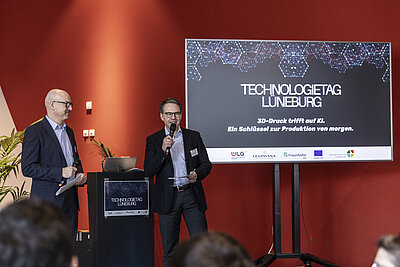 Zwei Männer stehen auf einer Bühne mit Mikrofonen und moderieren den Technologietag Lüneburg. Im Hintergrund eine Präsentationsfolie mit dem Titel "3D-Druck trifft auf KI. Ein Schlüssel zur Produktion von morgen." {f:if(condition:'Julia Crawley Photography',then:' | Bild: © Julia Crawley Photography',else:'')