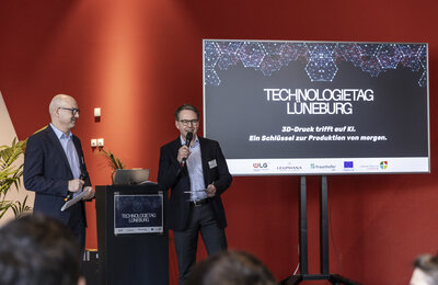 Zwei Männer stehen auf einer Bühne mit Mikrofonen und moderieren den Technologietag Lüneburg. Im Hintergrund eine Präsentationsfolie mit dem Titel "3D-Druck trifft auf KI. Ein Schlüssel zur Produktion von morgen." {f:if(condition:'Julia Crawley Photography',then:' | Bild: © Julia Crawley Photography',else:'')