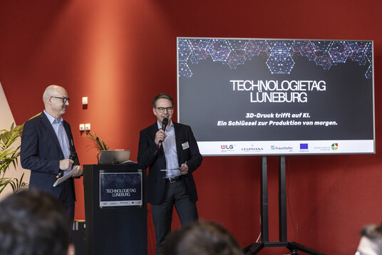 Moderation beim Technologietag Lüneburg | © Julia Crawley Photography Zwei Männer stehen auf einer Bühne mit Mikrofonen und moderieren den Technologietag Lüneburg. Im Hintergrund eine Präsentationsfolie mit dem Titel "3D-Druck trifft auf KI. Ein Schlüssel zur Produktion von morgen."