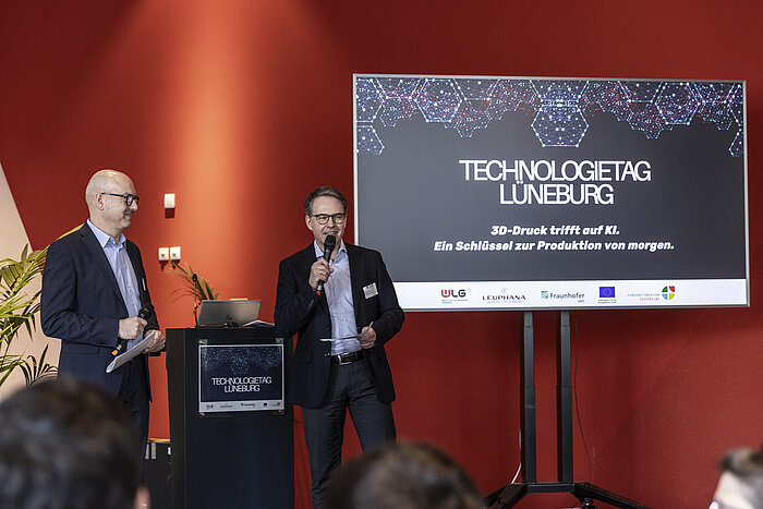 Zwei Männer stehen auf einer Bühne mit Mikrofonen und moderieren den Technologietag Lüneburg. Im Hintergrund eine Präsentationsfolie mit dem Titel "3D-Druck trifft auf KI. Ein Schlüssel zur Produktion von morgen." {f:if(condition:'Julia Crawley Photography',then:' | Bild: © Julia Crawley Photography',else:'')